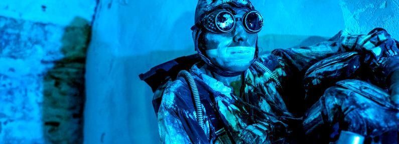 Un hombre con gafas y traje azul se sienta en un espacio iluminado de azul, mirando hacia adelante.