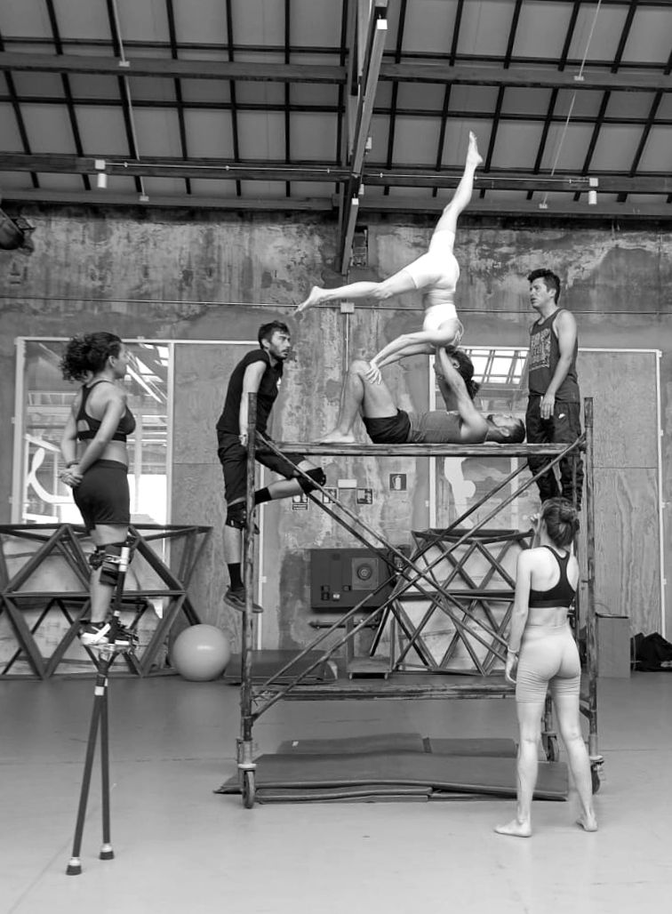 Acróbatas actuando sobre y alrededor de una estructura metálica en un gran estudio. Uno de ellos está de pie sobre zancos.