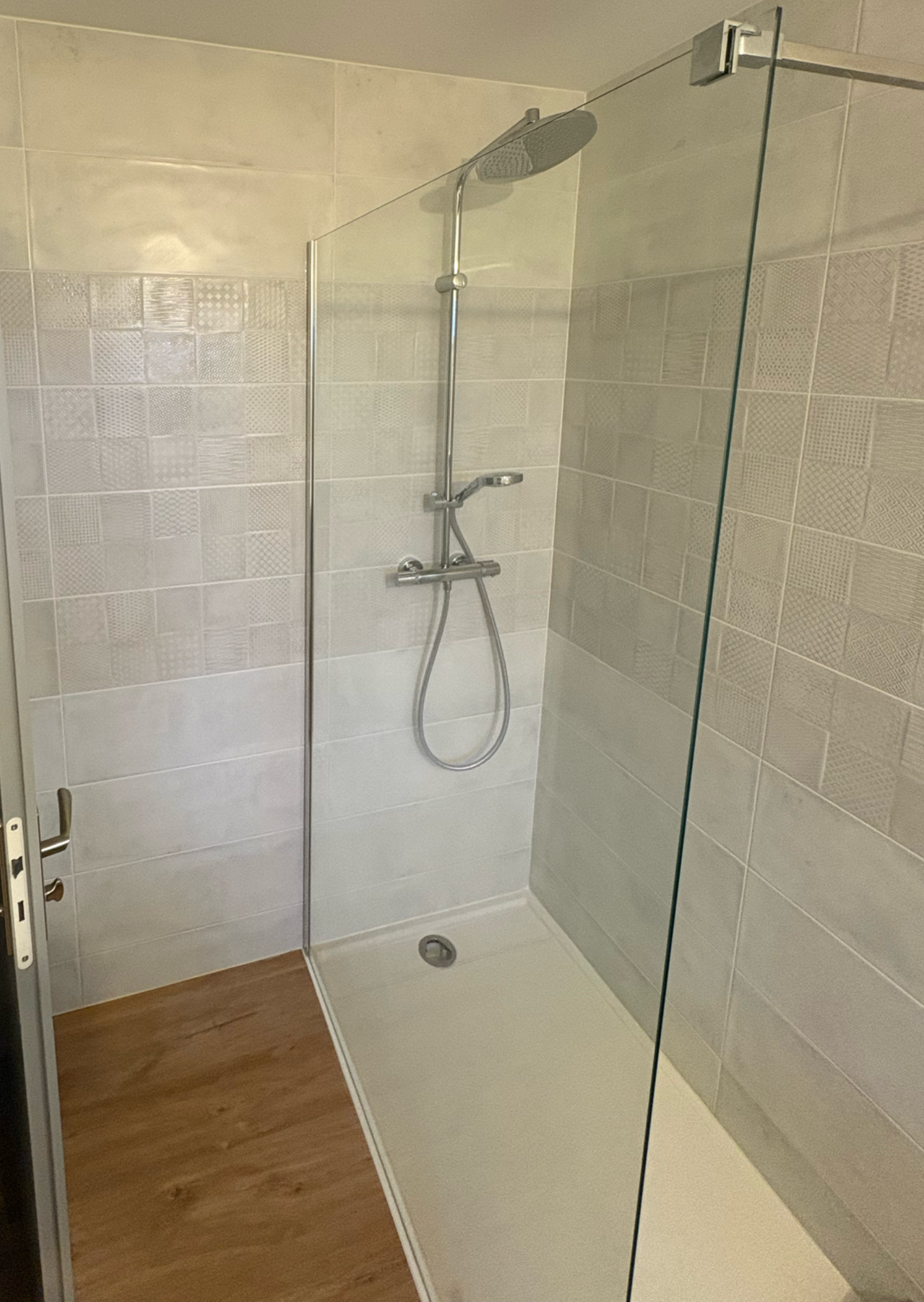 Douche à l'italienne moderne avec paroi en verre, murs carrelés, receveur blanc et sol imitation bois.