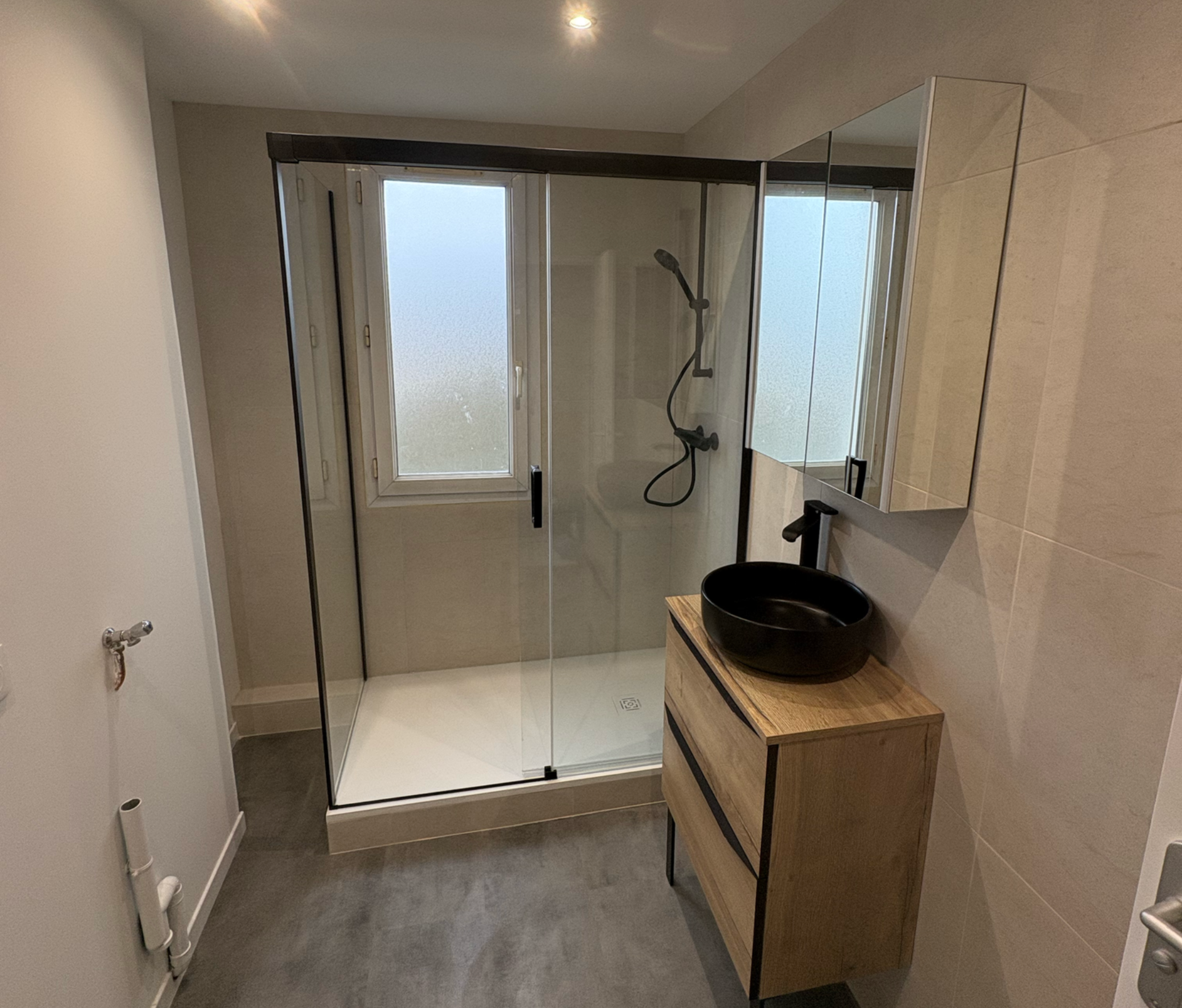 Salle de bains moderne avec douche en verre, fenêtre dépolie et meuble-lavabo en bois avec lavabo noir.