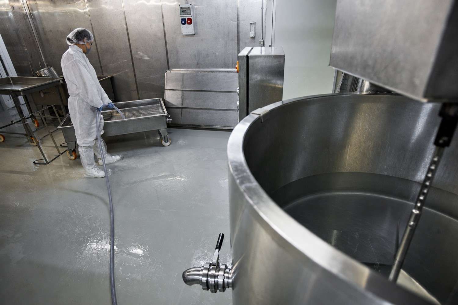 Trabajador con ropa protectora blanca de pie en una sala industrial de acero inoxidable con grandes tanques y suelo mojado.