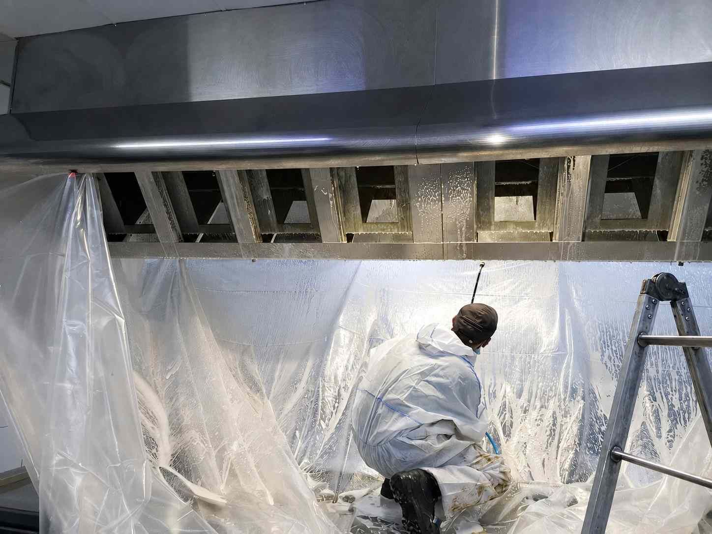 Un trabajador con traje protector pinta con aerosol una zona de ventilación del techo cubierta con una lámina de plástico.