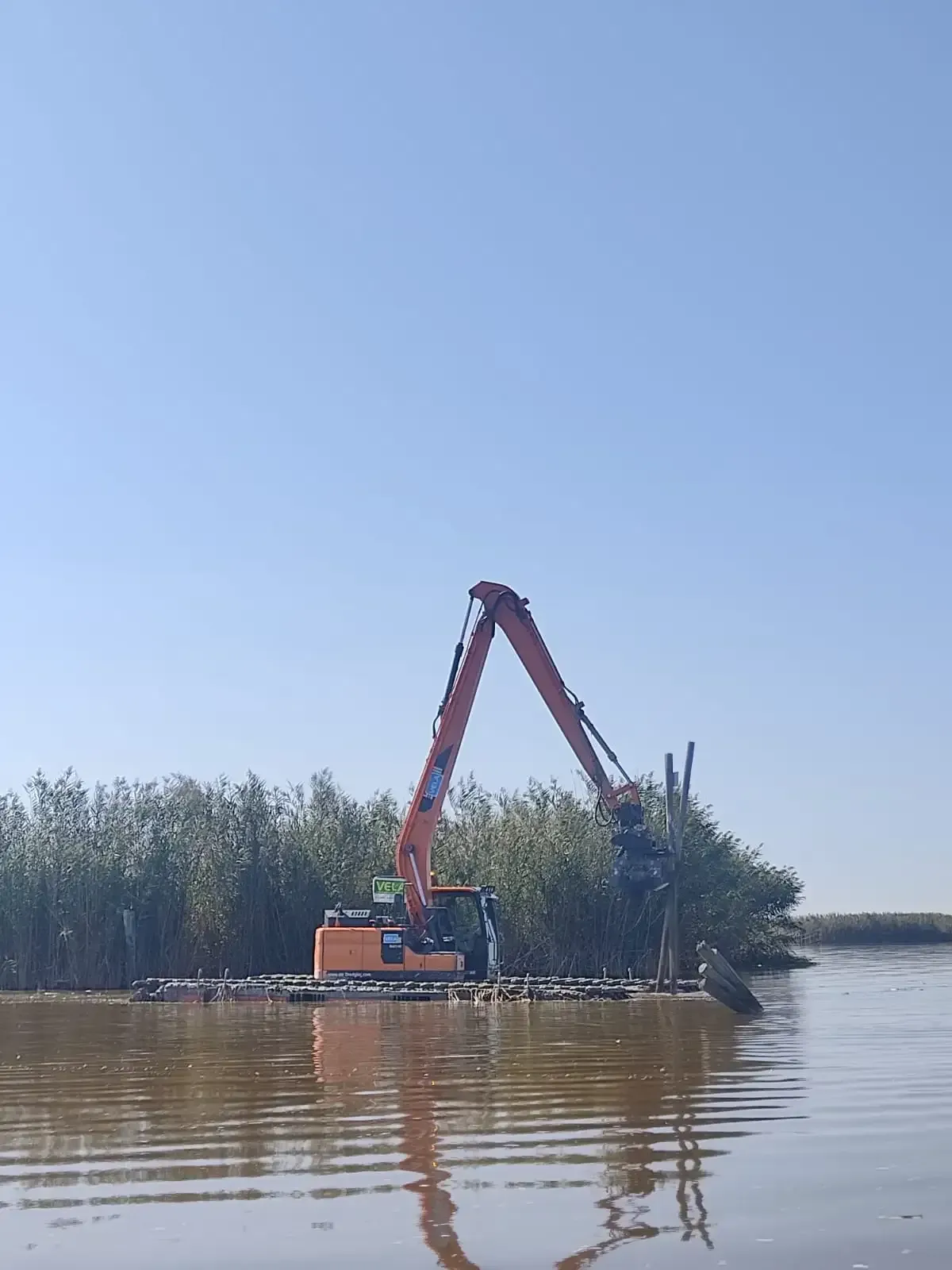 Excavadora naranja en una barcaza despejando juncos en un río tranquilo bajo un cielo azul despejado.