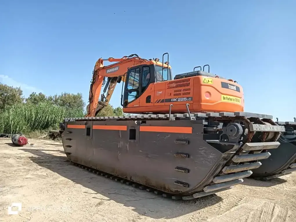 Excavadora naranja sobre grandes orugas metálicas en una obra de tierra bajo un cielo azul.