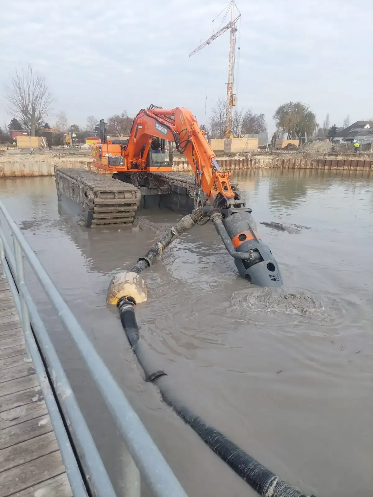 Una excavadora naranja draga agua turbia junto a un muelle, con una manguera flotante en primer plano.