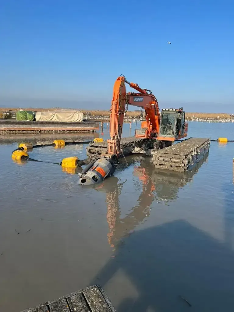 Una excavadora naranja trabaja en aguas poco profundas junto a la costa, moviendo una tubería sumergida.