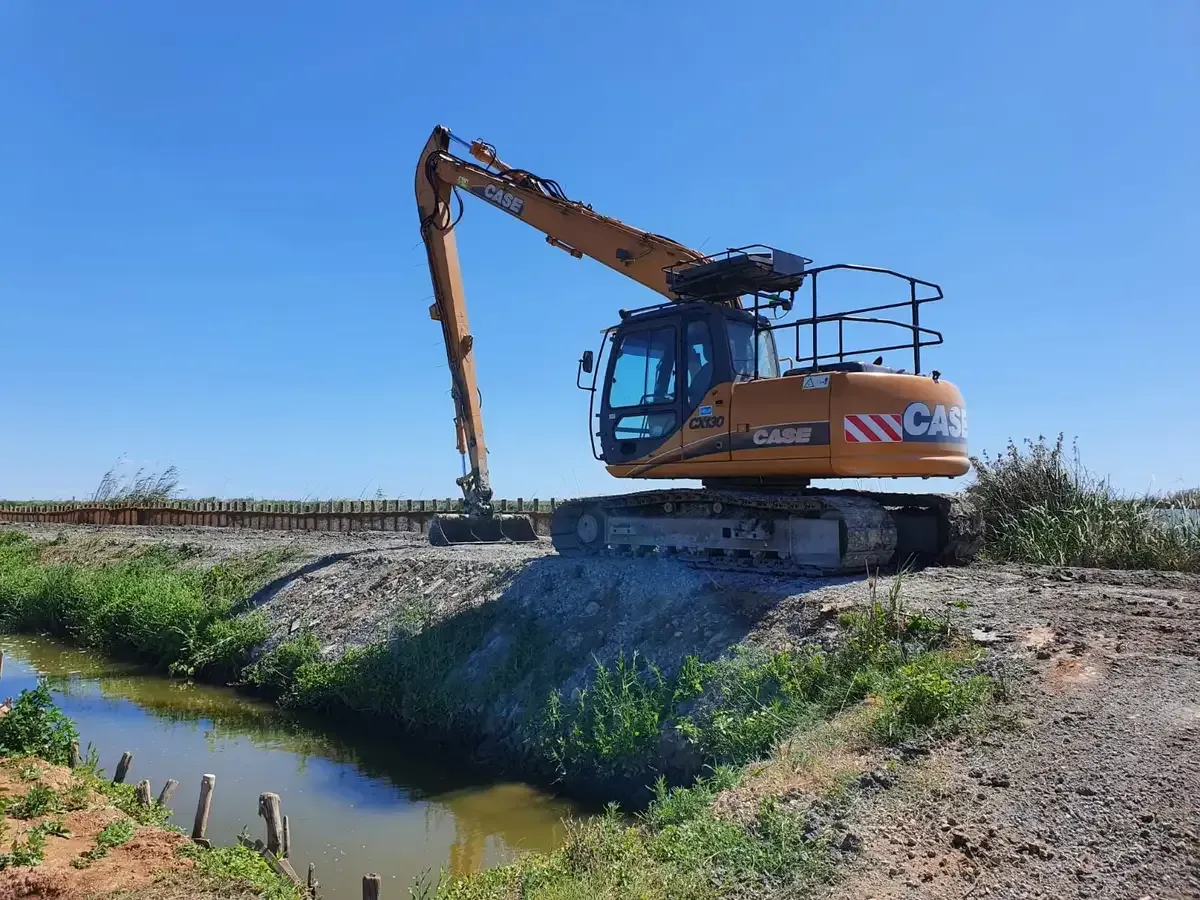 Una excavadora naranja excava junto a un estrecho canal de agua bajo un cielo azul despejado.