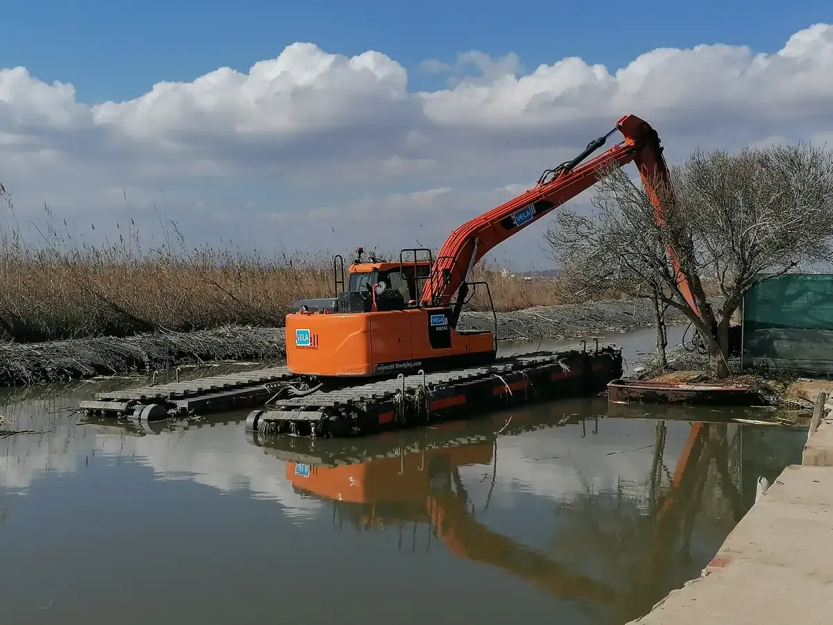 Excavadora y trabajadores con trajes de seguridad naranjas en un campo fangoso e inundado, cargando una pequeña embarcación.