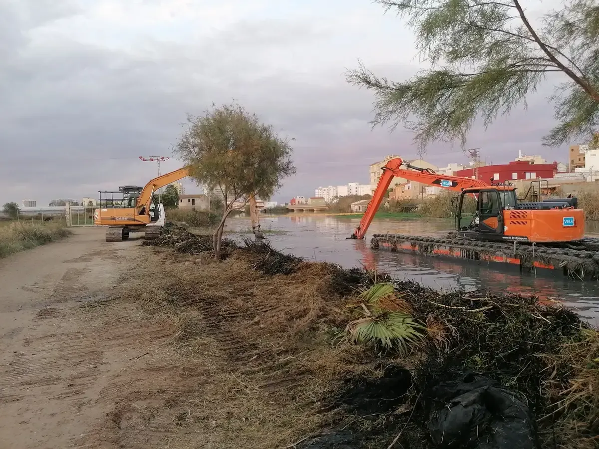 Excavadoras dragando un canal fangoso junto a un camino de tierra bajo un cielo nublado.