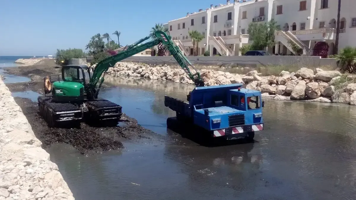 Una excavadora carga lodo en un camión volquete azul junto a los edificios de la ribera de un canal poco profundo.