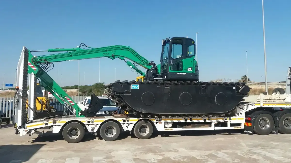Excavadora verde cargada en un remolque de plataforma, siendo transportada por una carretera soleada.