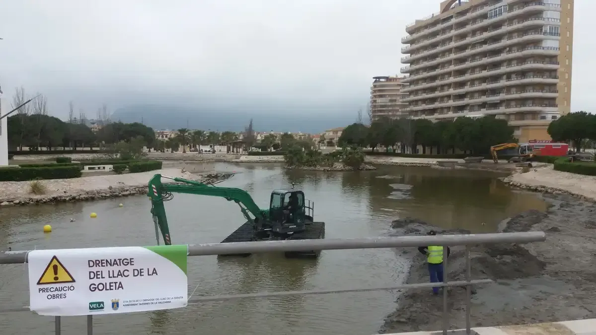 Ribera inundada con una pequeña excavadora, una señal de advertencia y un edificio de apartamentos alto al fondo.