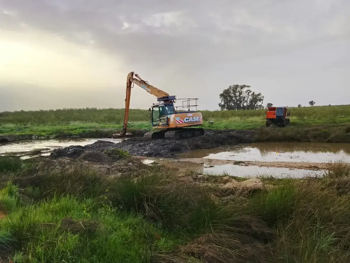 Obreros de la construcción con chalecos naranjas inspeccionan una excavadora que está removiendo tierra en una obra.