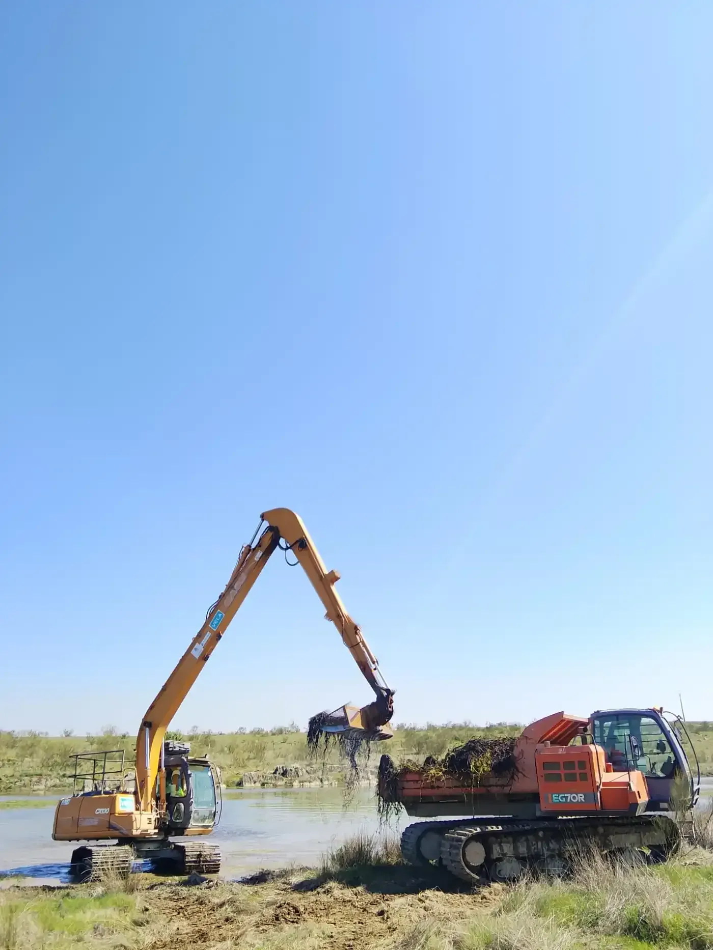 Unos trabajadores manejan una retroexcavadora junto a un río, entre campos verdes y rocas.