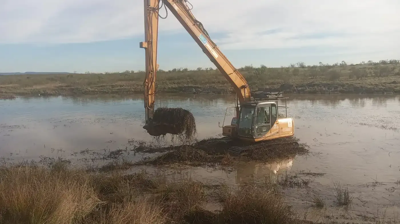 Trabajadores con chalecos de seguridad y cascos inspeccionan una excavadora amarilla en una obra de construcción fangosa a orillas del río.