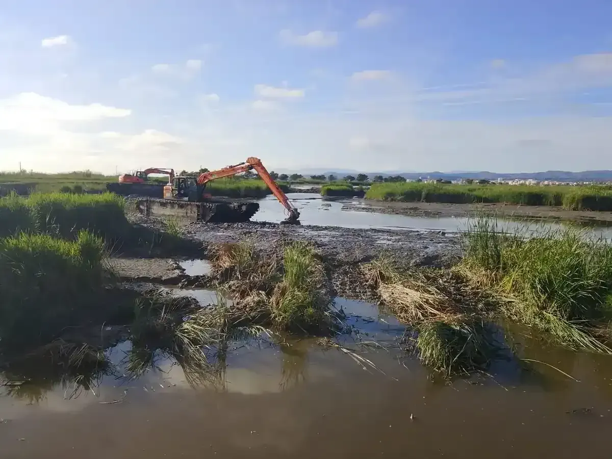 Excavadora y trabajadores con trajes de seguridad naranjas en un campo fangoso e inundado, cargando una pequeña embarcación.