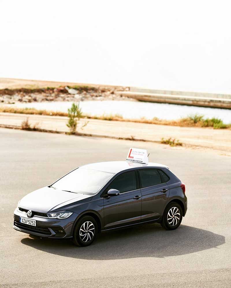 Γκρι Volkswagen Polo με πινακίδα «μαθητής οδήγησης» στην οροφή του σε πλακόστρωτη περιοχή κοντά σε υδάτινο σώμα.