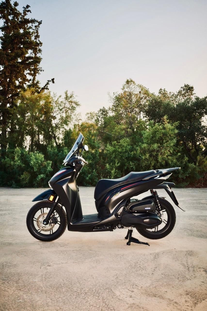 Μαύρο σκούτερ παρκαρισμένο σε χαλίκι με δέντρα στο φόντο.