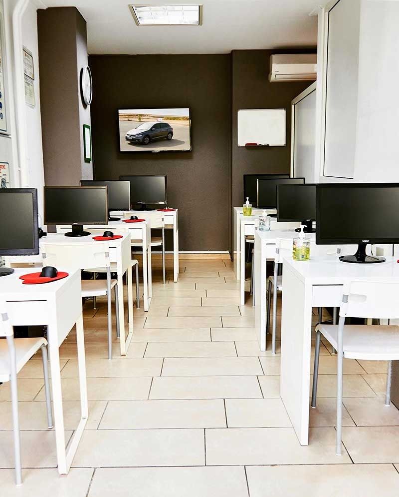 Σειρές από λευκά θρανία με υπολογιστές σε μια τάξη· μια τηλεόραση δείχνει ένα αυτοκίνητο, με σκούρο καφέ τοίχο και πλακάκια στο δάπεδο.