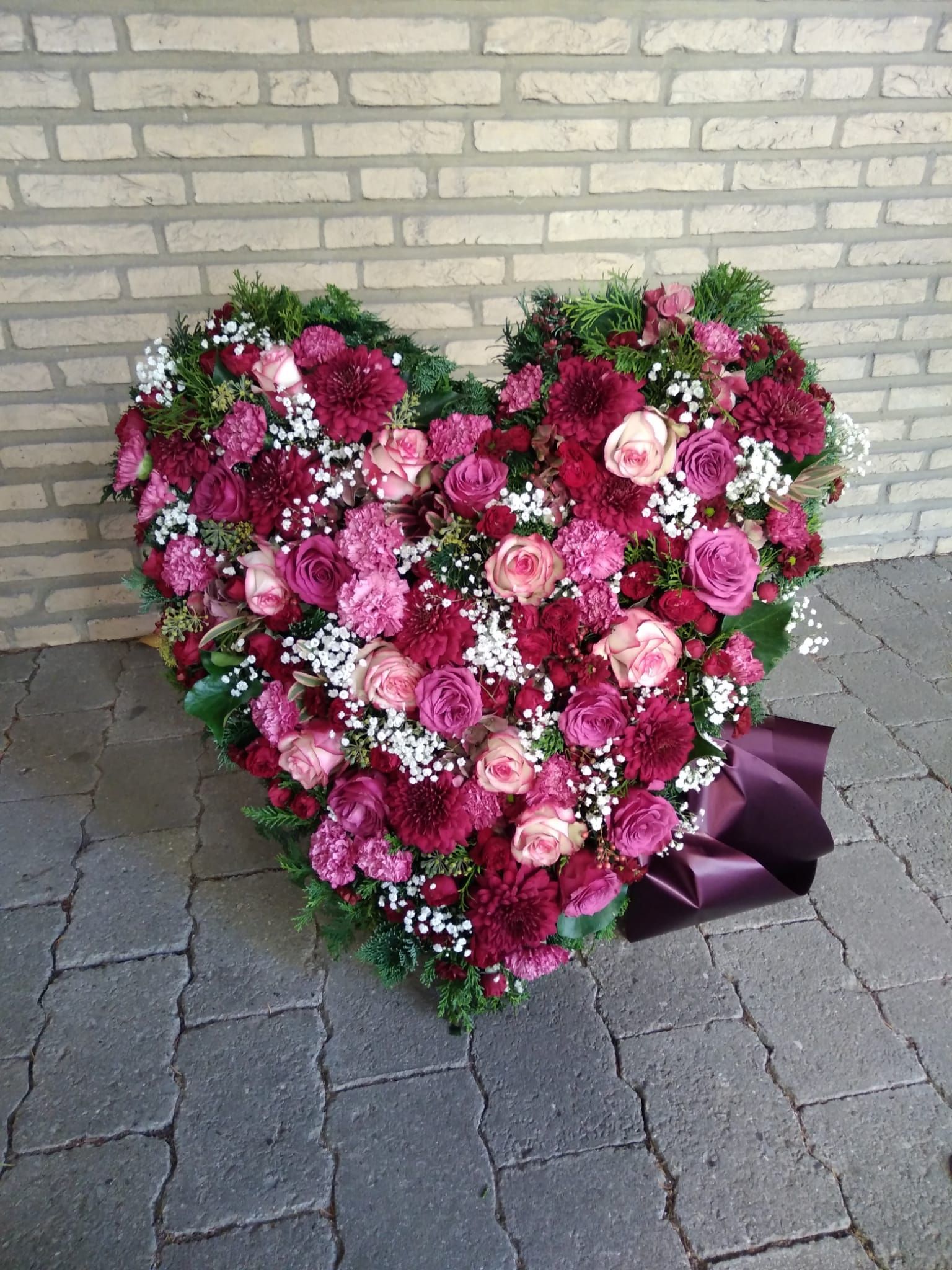 Ein herzförmiger Kranz aus rosa und violetten Blumen liegt auf einem Gehweg vor einer Backsteinmauer.