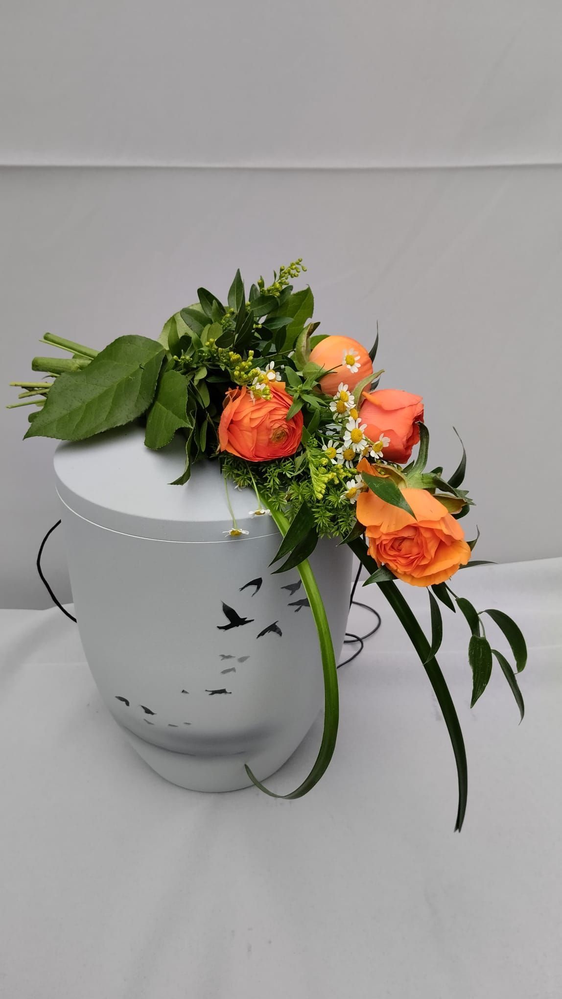 Eine weiße Urne gefüllt mit orangefarbenen Rosen und Grünpflanzen auf einem Tisch.