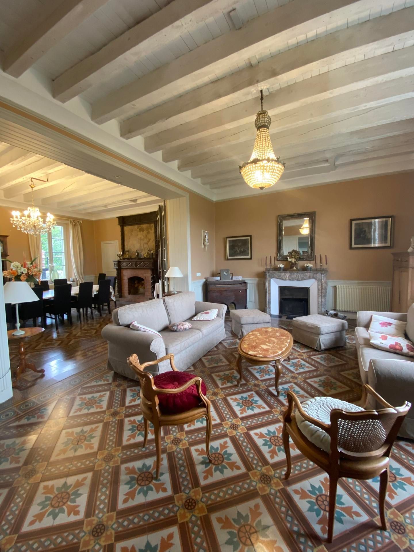 Salon spacieux avec sol carrelé à motifs, poutres apparentes et plusieurs espaces de détente agrémentés d'un lustre.