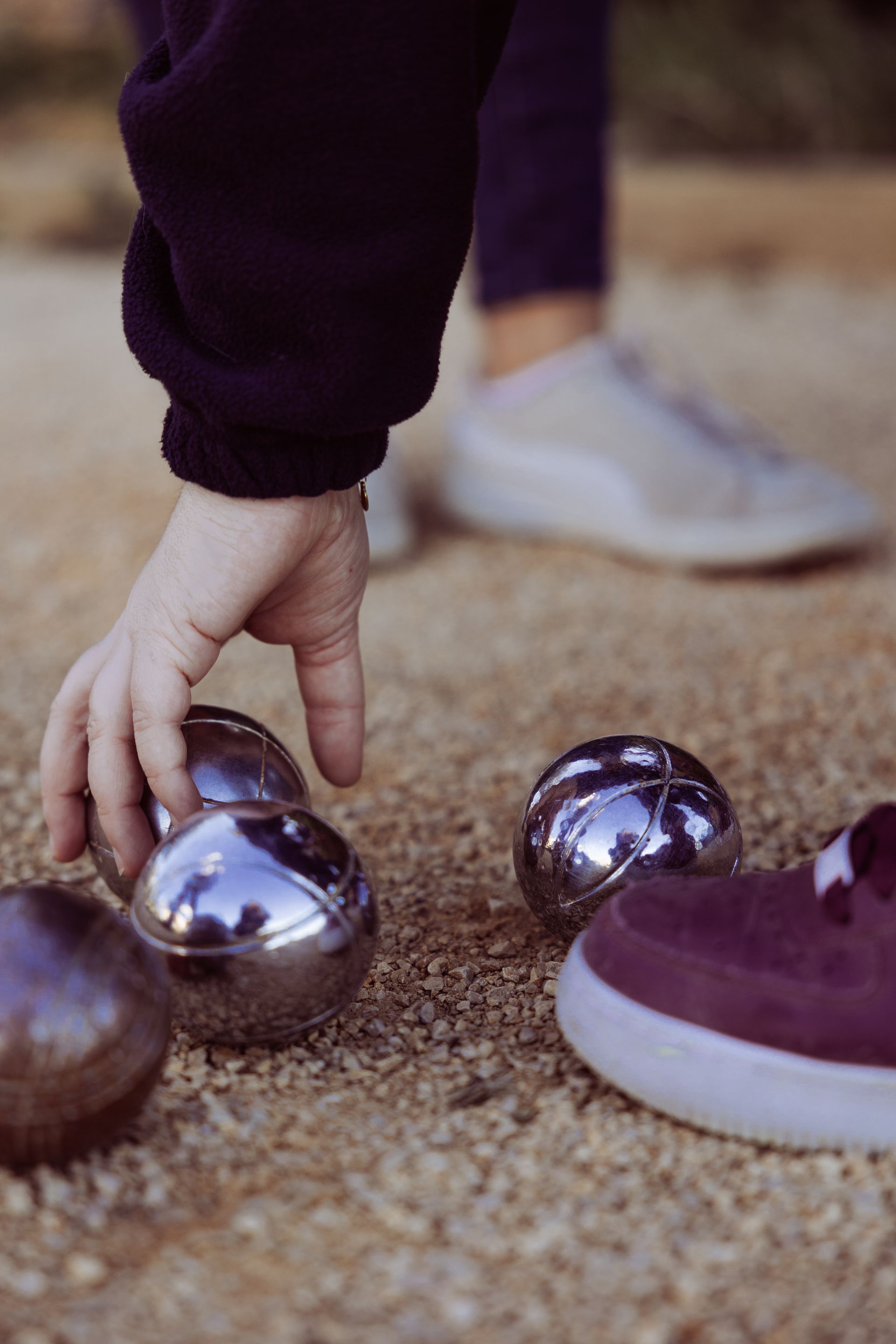La main d'une personne tend la main vers des boules de métal brillantes posées sur une surface de gravier, près d'un pied chaussé.
