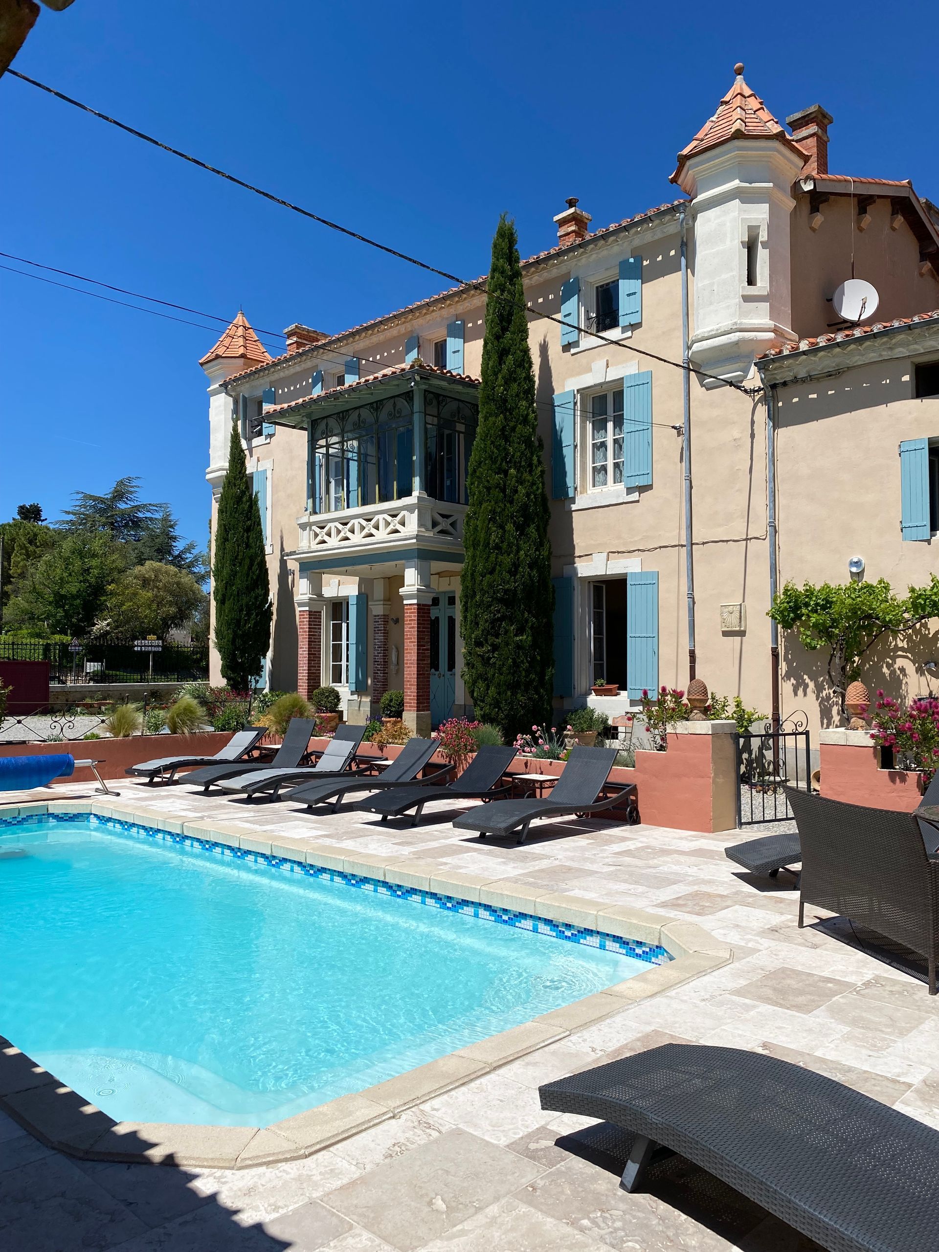Extérieur d'une villa avec piscine, grands arbres et volets bleus par une journée ensoleillée.