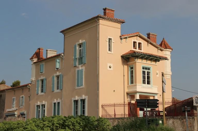 Bâtiment beige de trois étages avec des volets bleus et un toit de tuiles rouges, sous un ciel bleu clair.