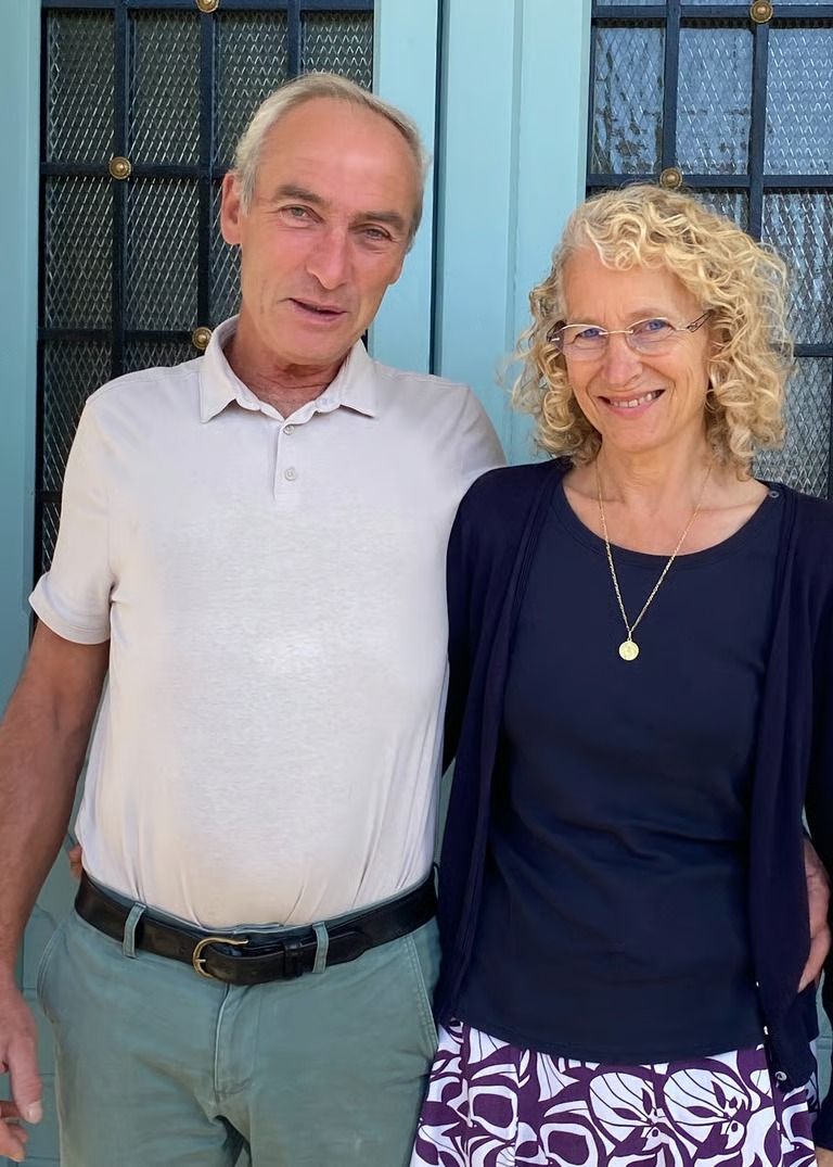 Un homme et une femme se tiennent côte à côte devant une porte bleue. La femme porte des lunettes et un gilet, et sourit. L'homme a un bras autour d'elle.