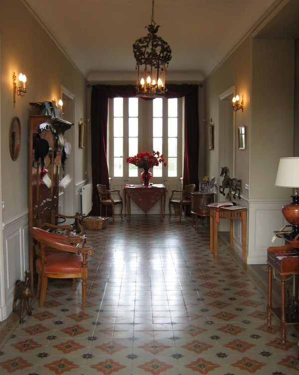 Long couloir avec sol carrelé à motifs, meubles et grande fenêtre avec rideaux rouges.