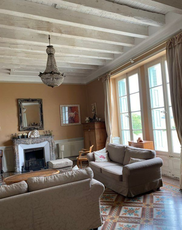 Salon avec murs beiges, poutres blanches au plafond, lustre, cheminée et deux canapés beiges.