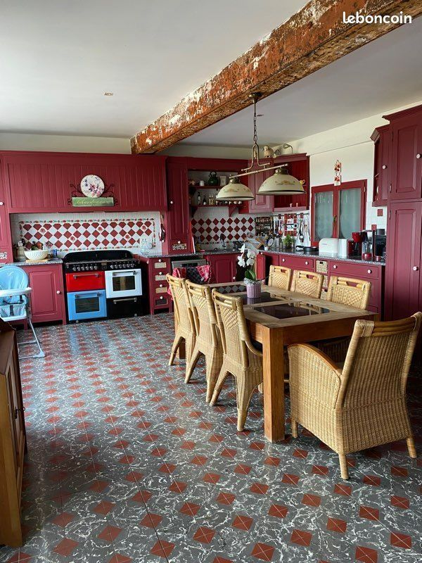 Cuisine avec placards rouges, sol carrelé, table à manger, cuisinière et poutre apparente.