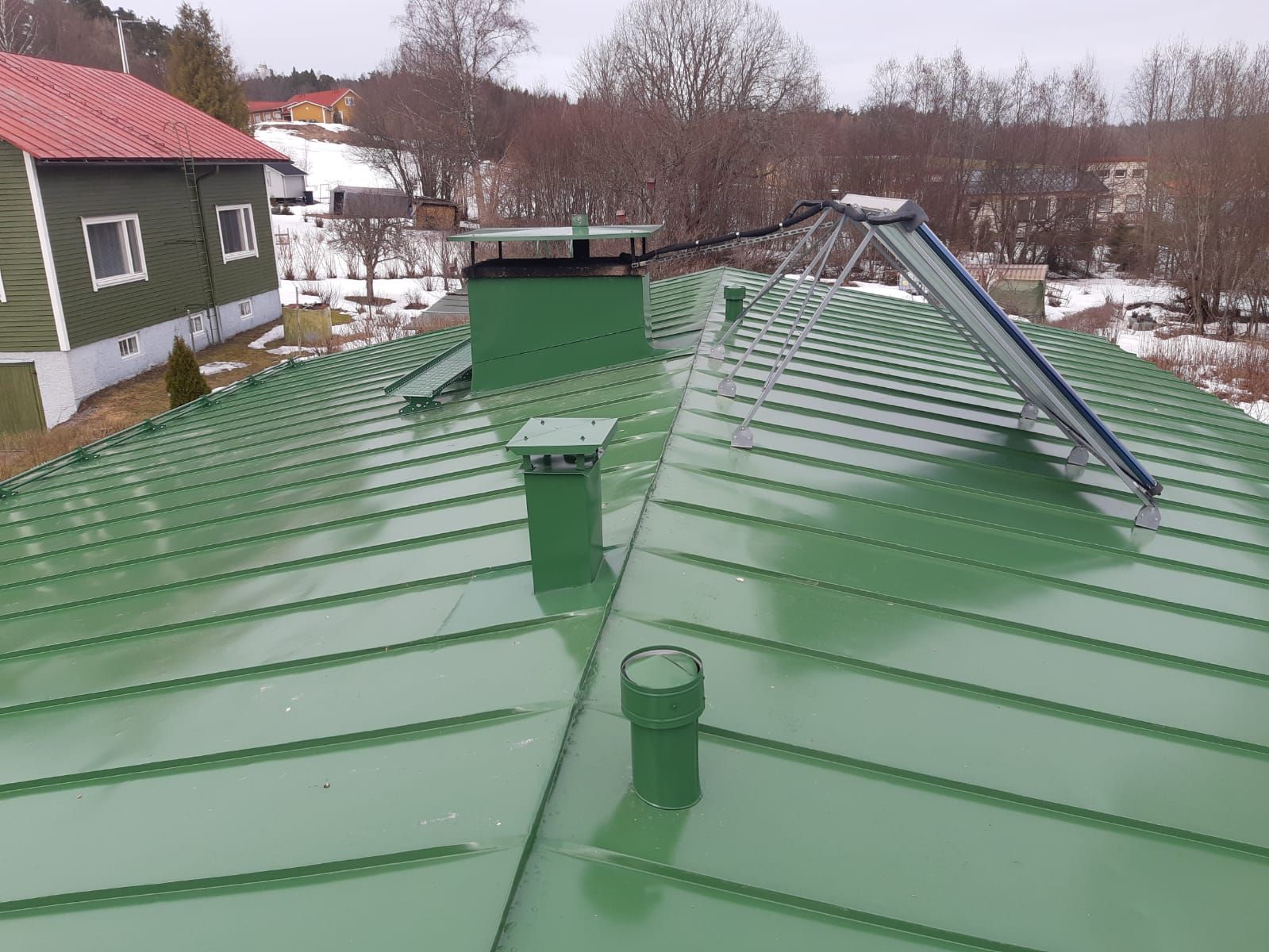 Vihreä peltikatto, jossa on savupiippu, tuuletusaukot ja aurinkopaneeli, taustalla talo ja lunta.