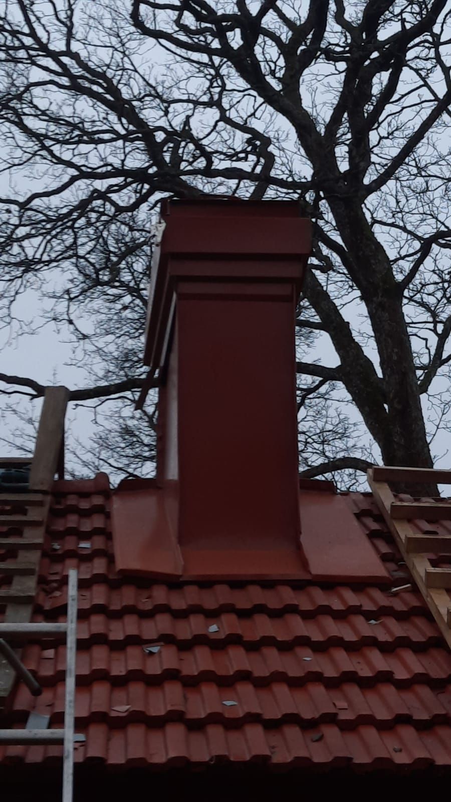 Punainen savupiippu punaisella tiilikatolla, taustalla paljas puu.