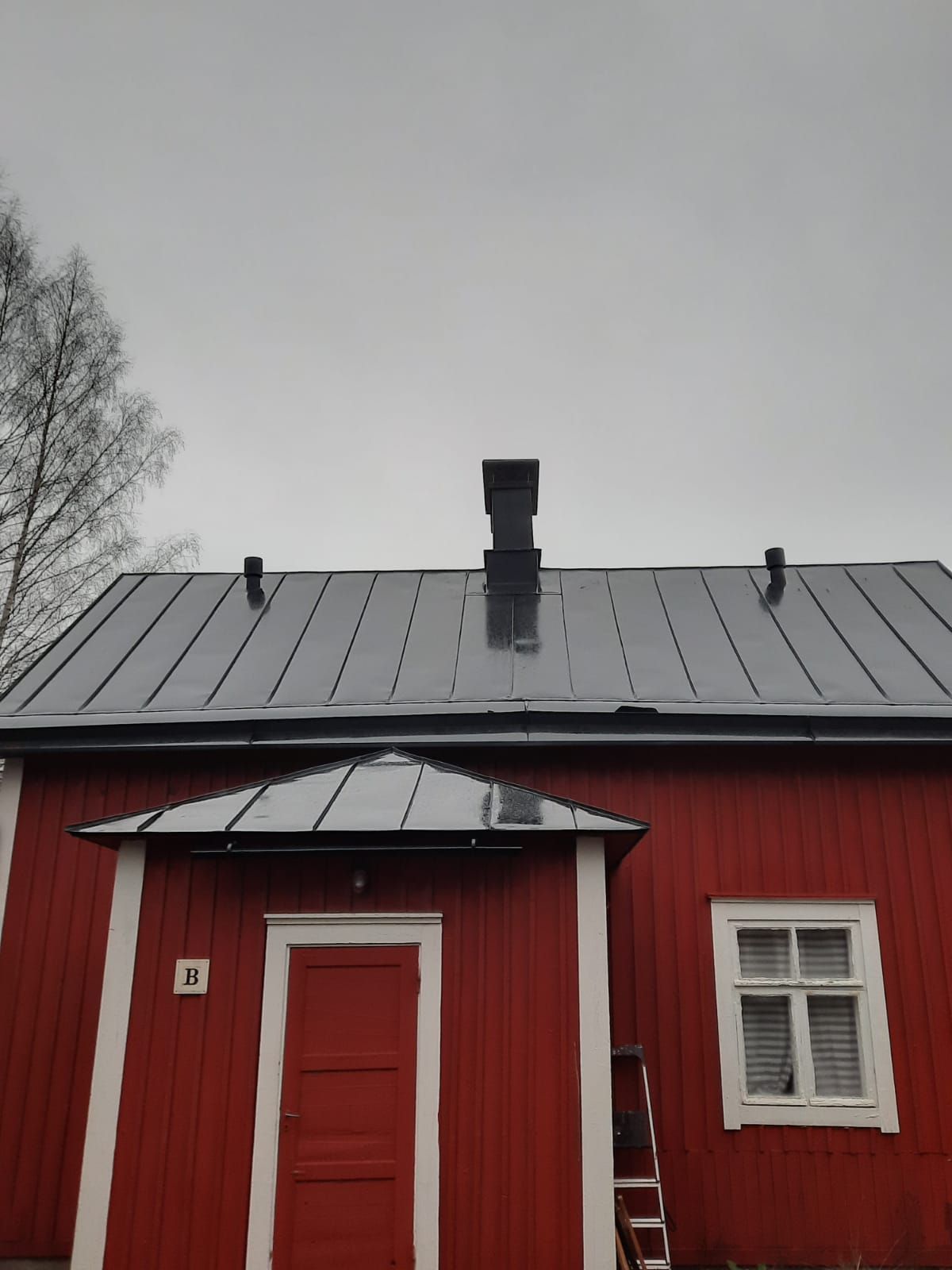 Punainen puinen rakennus, jossa on harmaa peltikatto, savupiippu ja pieni etukuisti.