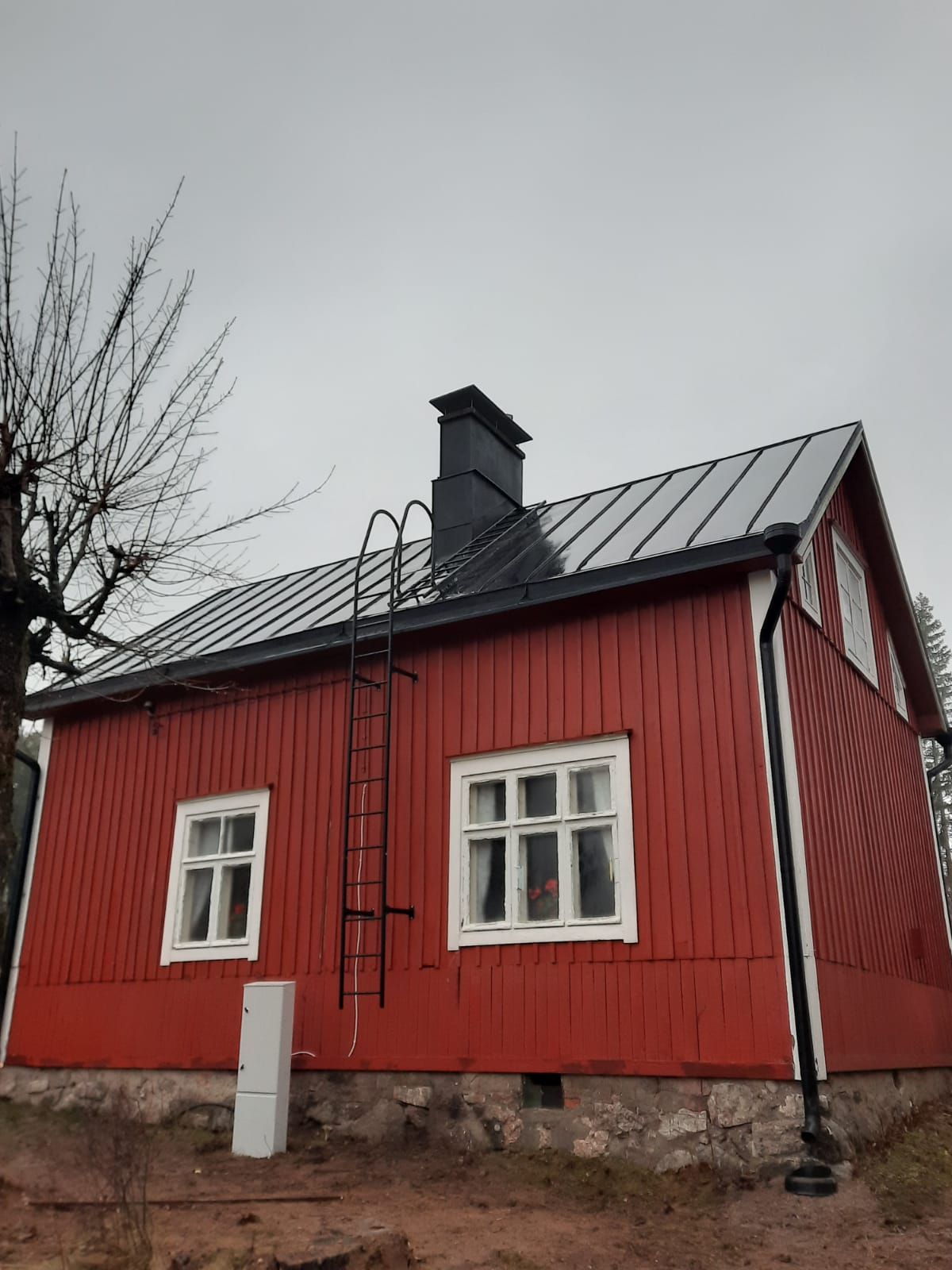 Punainen puutalo mustalla metallikatolla, tikkailla ja savupiipulla pilvisen taivaan alla.