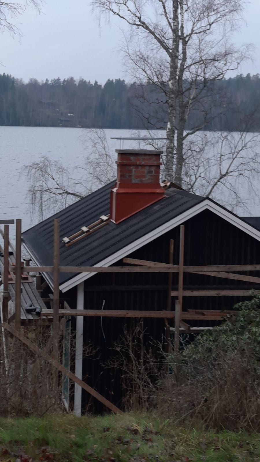 Mustakattoinen mökki, jossa on punainen savupiippu, rakennustelineet ja järvi taustalla.