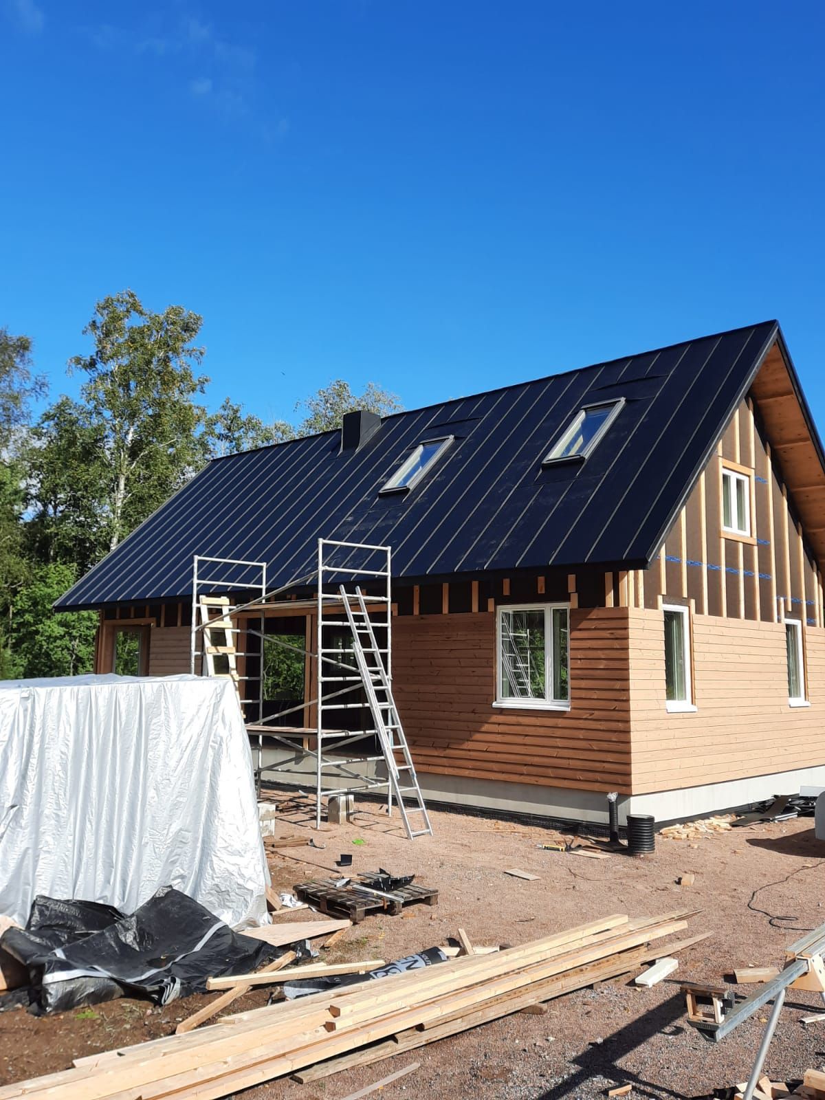 Rakenteilla oleva talo mustalla katolla, puisilla ulkoverhouksilla, sinisellä taivaalla.