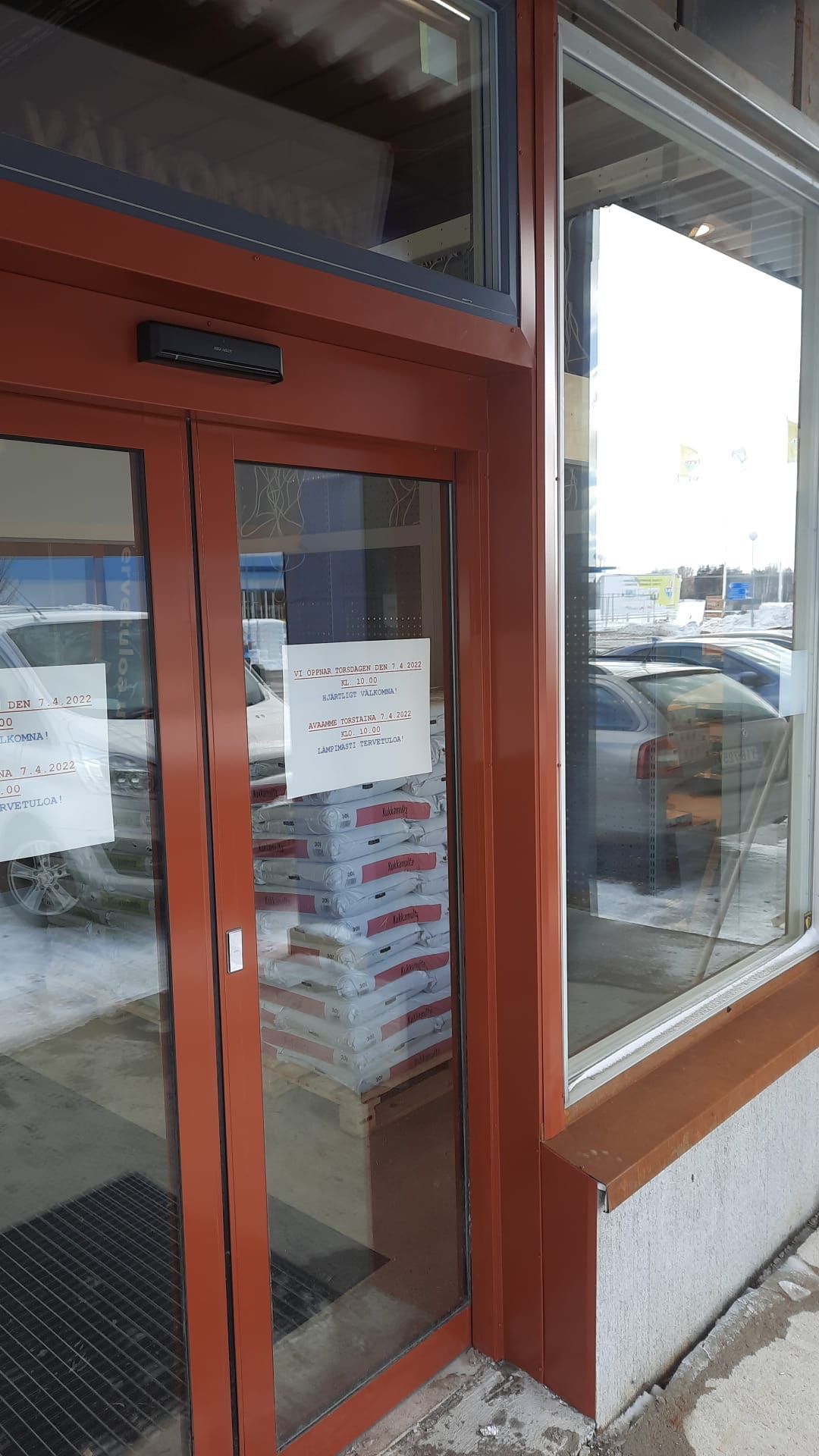 Punakehyksinen lasiovi liikkeessä, osittain paperipinojen peitossa. Ovessa näkyy kyltti.