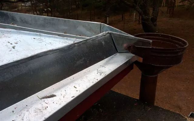 Sinkitty metallinen ränni, joka tyhjenee ruostuneeseen, pyöreään säiliöön. Ulkokuva punaiselta katolta.