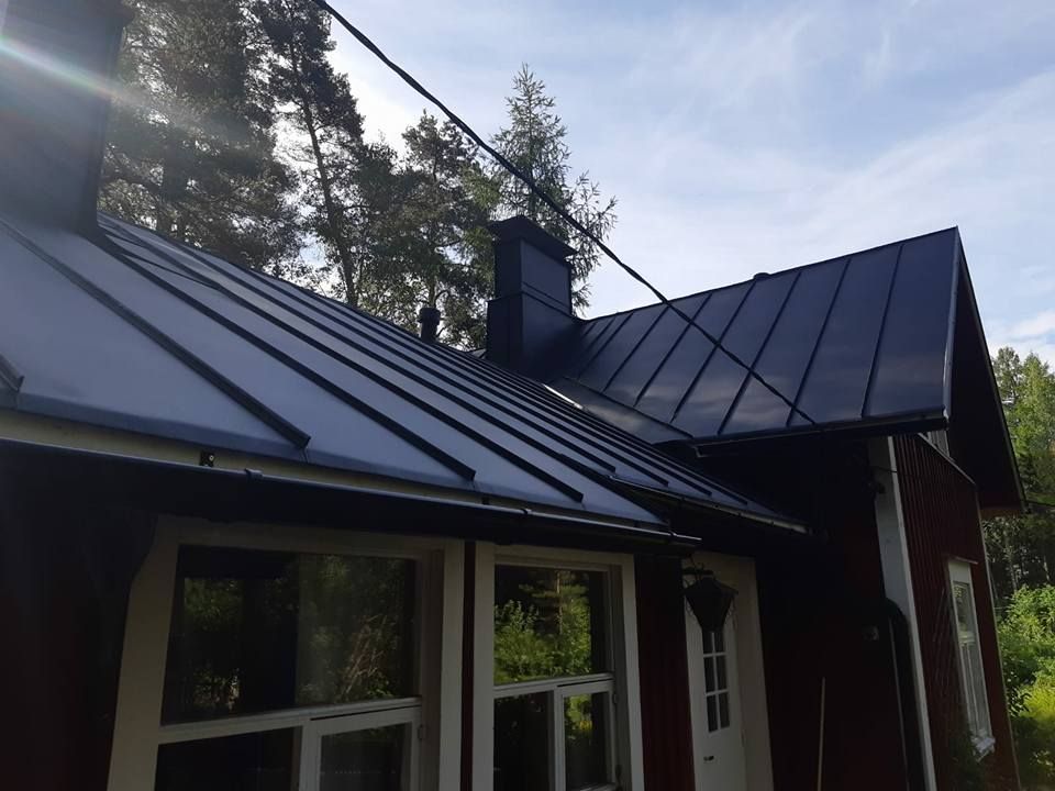 Punainen talo, jossa on musta metallikatto, savupiiput, ikkunat ja sähkölinjat. Taustalla puita.