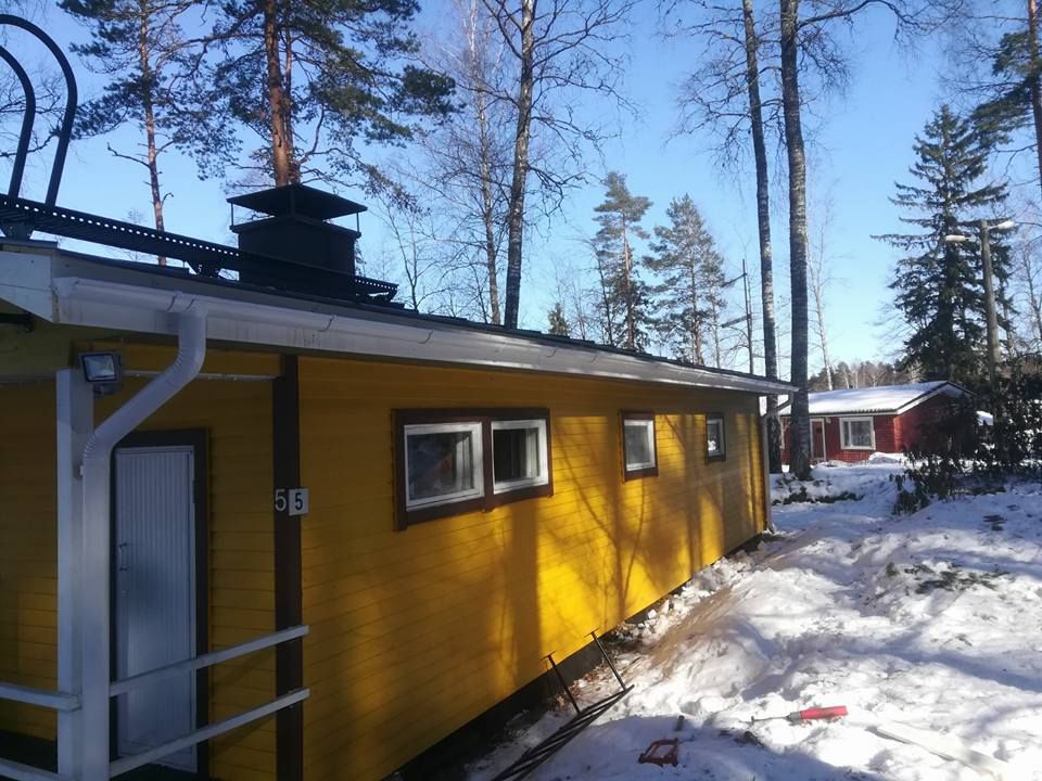 Keltainen mökki valkoisilla reunuksilla, luminen maa, puita taustalla, toinen punainen mökki näkyvissä.