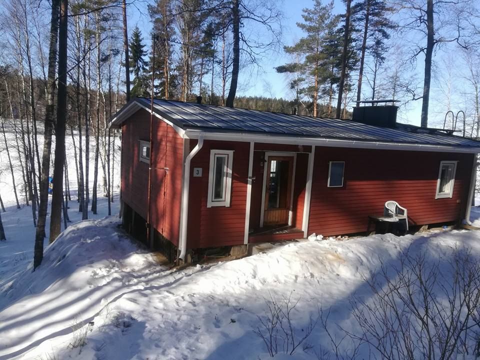 Punainen puinen mökki lumimaisemassa, puiden ympäröimänä kirkkaan taivaan alla.
