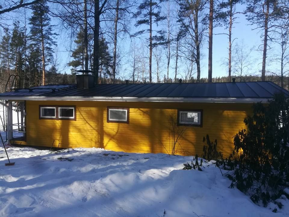 Keltainen mökki tummalla katolla lumimaisemassa, puiden ympäröimänä sinisen taivaan alla.
