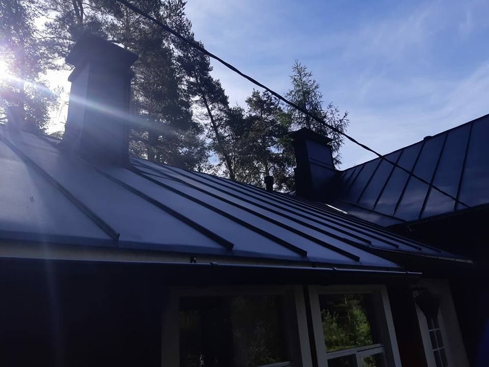 Musta katto kahdella savupiipulla aurinkoista taivasta vasten, puita taustalla.
