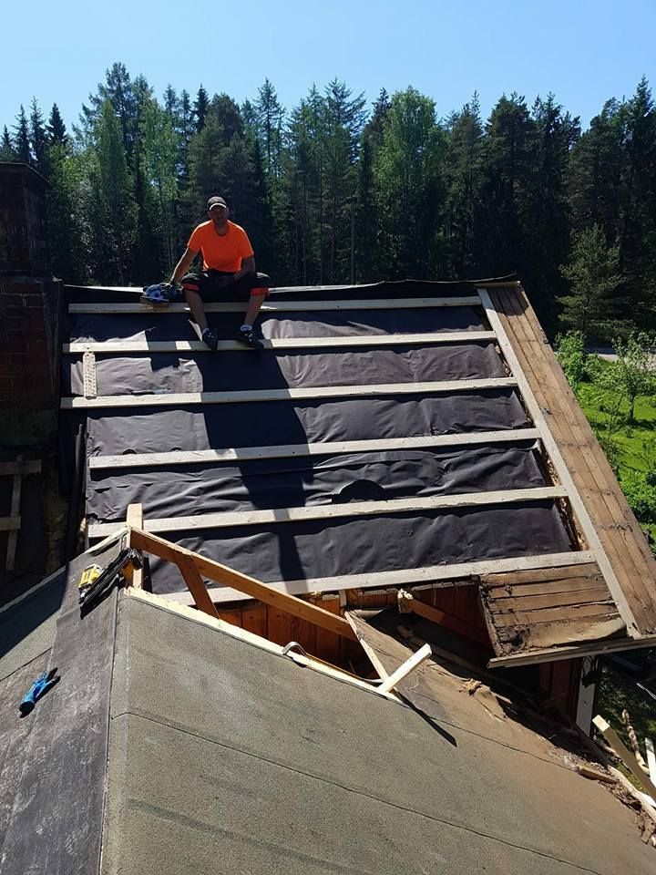 Mies istuu rakenteilla olevalla katolla yllään oranssi paita, musta aluskate ja puiset tuet näkyvissä.