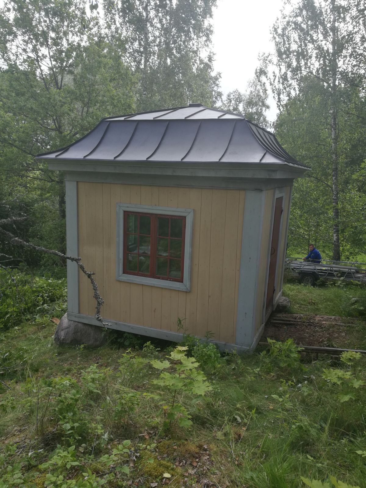 Pieni, keltainen rakennus tummalla katolla metsäisellä alueella; punaisella reunustettu ikkuna.