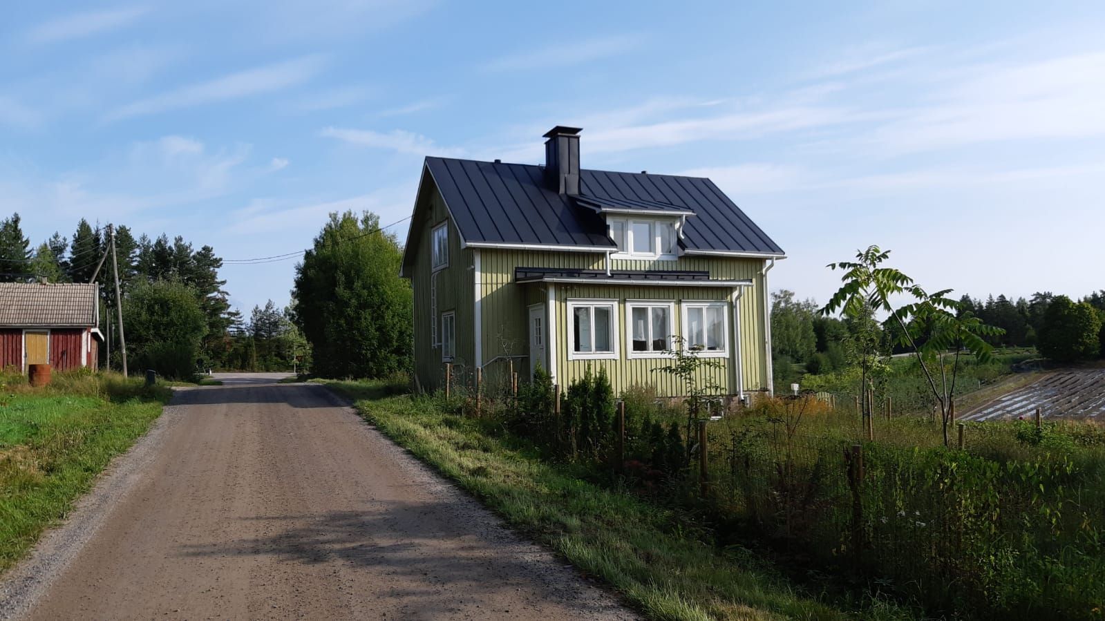 Keltainen, tummakattoinen talo seisoo hiekkatien vieressä sinisen taivaan alla.