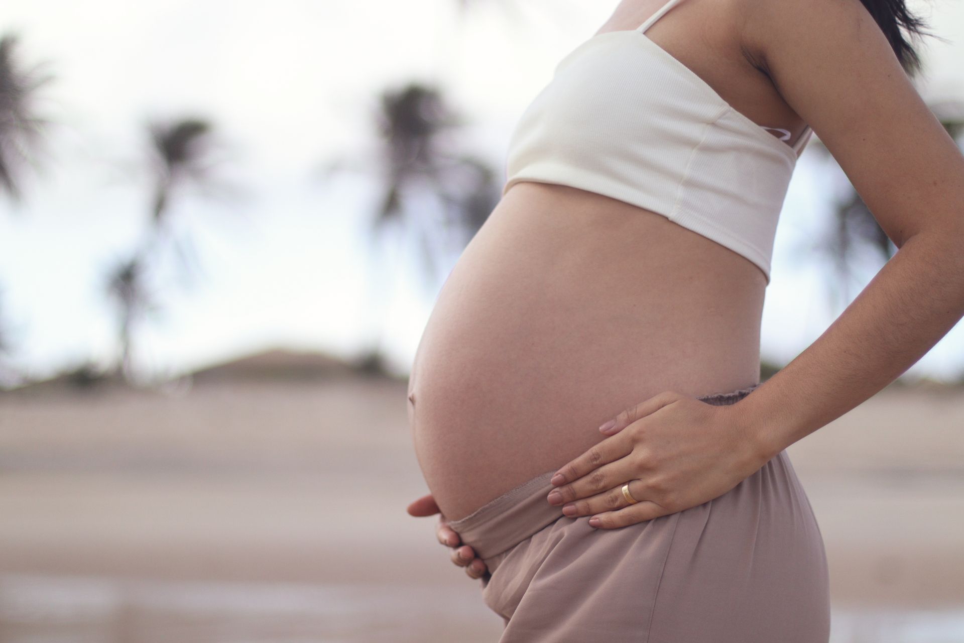 Une femme enceinte se tient le ventre sur une plage, avec des palmiers flous en arrière-plan.